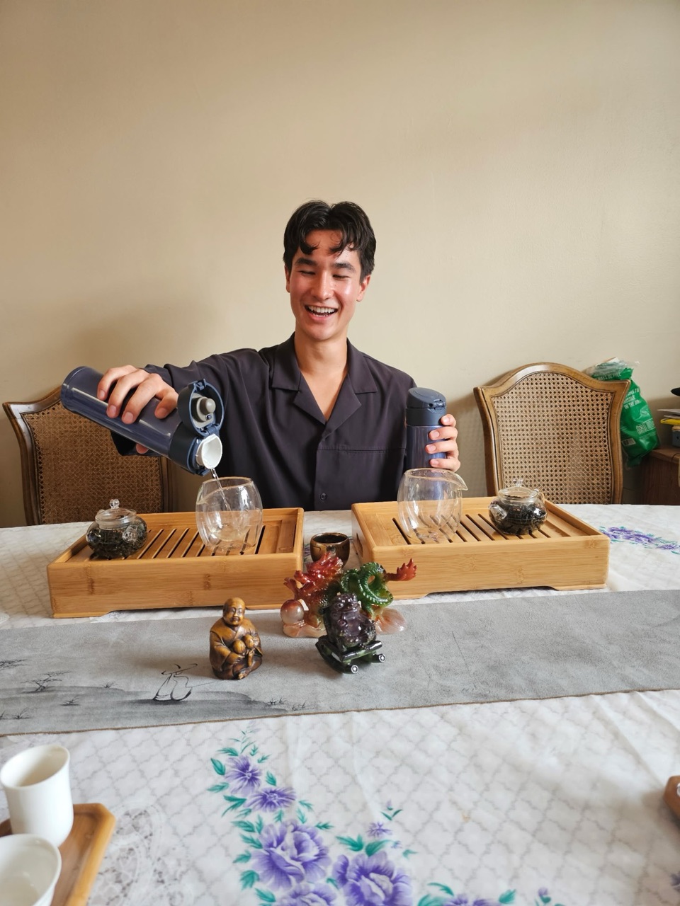 Ethan laughing as he pours hot water for gong fu tea at a table set with bamboo tea trays and small glass teapots