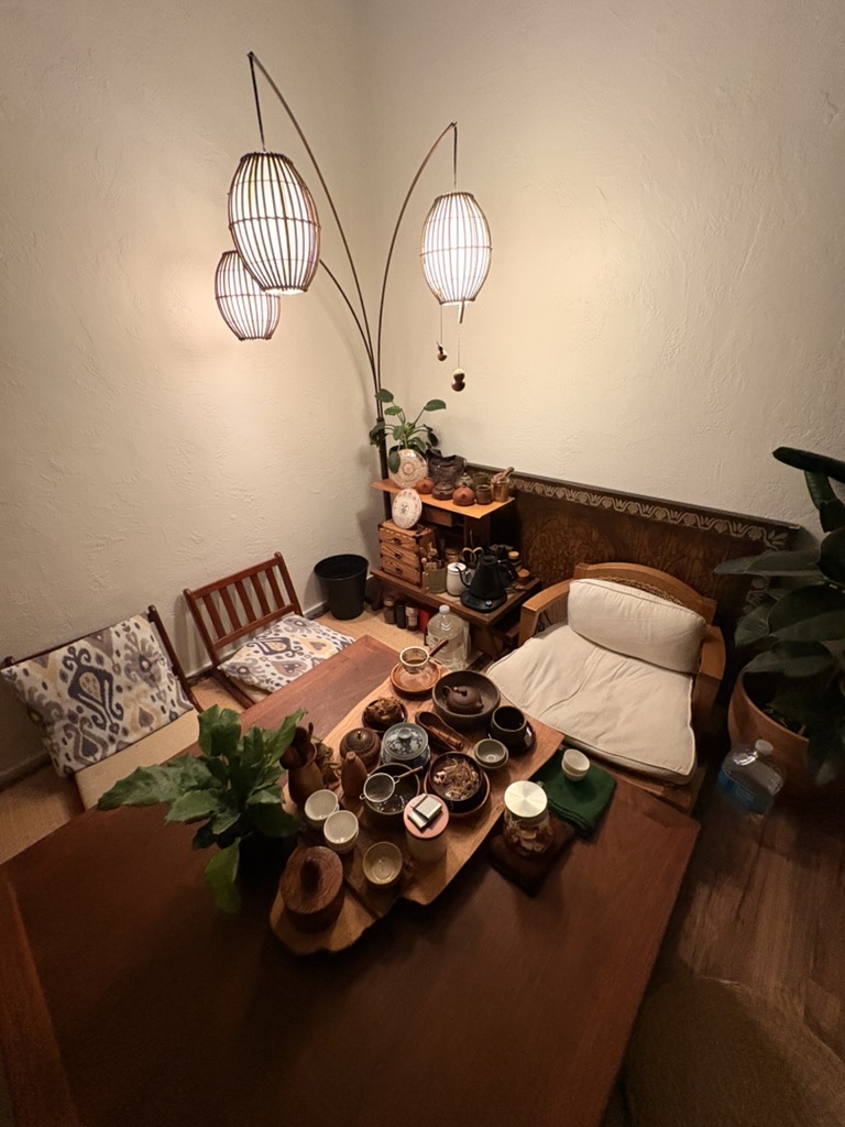 The Tea Temple: a small lamp-lit room with woven pendant lamps, a wooden tea table covered in teapots and cups, a cushioned chair, floor pillows, and plants